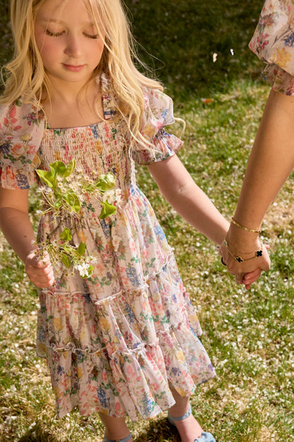 A young girl with long blonde hair in Ivy City Co's Mini Madeline Dress in Cordelia Floral holds wildflowers and an adult's hand while standing on a sunlit grassy lawn.