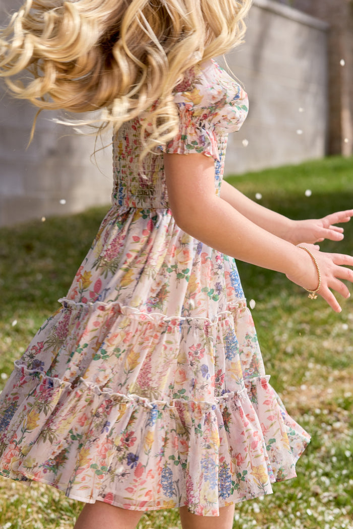 A young girl with long, wavy blonde hair twirls outside in the Ivy City Co Mini Madeline Dress in Cordelia Floral. Sunlight catches her flowing hair and skirt as she stands on grass by a concrete wall with white petals swirling around her.