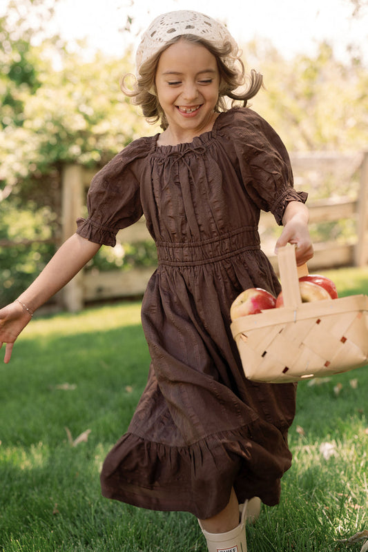 A young girl wearing the Ivy City Co Mini Maleah Dress in Warm Cocoa, complete with charming puff sleeves and a white hat, smiles as she runs across the grass with a basket of apples in a sunny outdoor setting.
