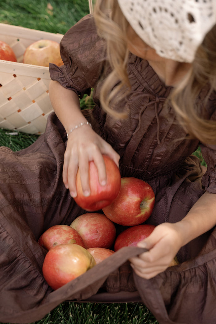 A child wearing the Ivy City Co Mini Maleah Dress in Warm Cocoa, featuring charming puff sleeves, and a white hat sits on grass holding several red apples in her lap, with a basket of apples beside her.