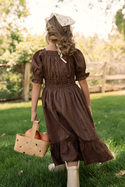 A young girl wearing the Ivy City Co Mini Maleah Dress in Warm Cocoa with puff sleeves and boots walks on grass, holding a basket of apples. She has wavy hair styled with a white lace headscarf, and a wooden fence and trees can be seen in the background.