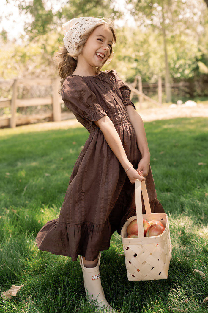 A smiling young girl wears the Mini Maleah Dress in Warm Cocoa by Ivy City Co, featuring charming puff sleeves, and pairs it with white boots as she holds a basket of apples outdoors on green grass. The soft viscose fabric of her brown dress enhances both comfort and style against a backdrop of trees and a wooden fence.