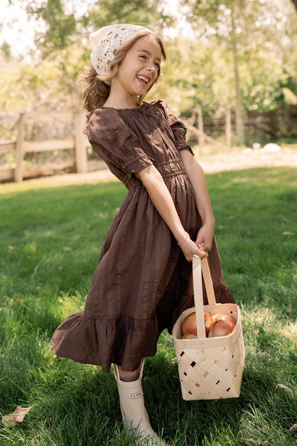 A smiling young girl wears the Mini Maleah Dress in Warm Cocoa by Ivy City Co, featuring charming puff sleeves, and pairs it with white boots as she holds a basket of apples outdoors on green grass. The soft viscose fabric of her brown dress enhances both comfort and style against a backdrop of trees and a wooden fence.