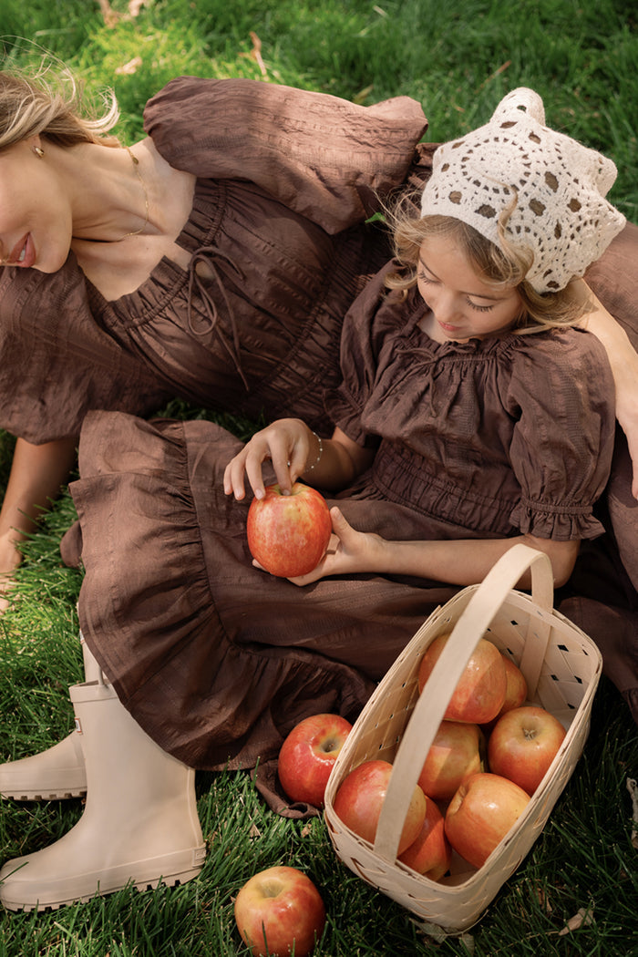 A woman and a girl sit on the grass holding apples, with a basket of apples beside them. The girl wears a white crocheted bonnet and is dressed in the charming Mini Maleah Dress in Warm Cocoa by Ivy City Co, crafted from soft viscose.