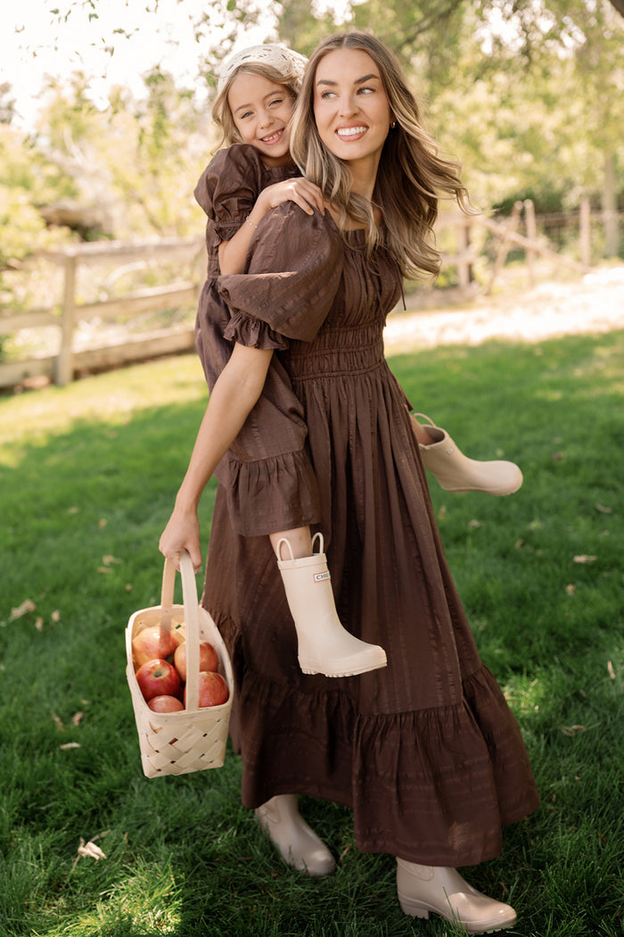 A woman wearing the Ivy City Co Mini Maleah Dress in Warm Cocoa with puff sleeves carries a smiling girl on her back. Both are dressed in matching Mini Maleah Dresses paired with boots. The woman holds a basket of apples as they stand together outdoors on green grass near a wooden fence.