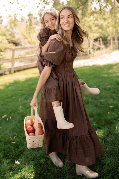 A woman wearing the Ivy City Co Mini Maleah Dress in Warm Cocoa with puff sleeves carries a smiling girl on her back. Both are dressed in matching Mini Maleah Dresses paired with boots. The woman holds a basket of apples as they stand together outdoors on green grass near a wooden fence.