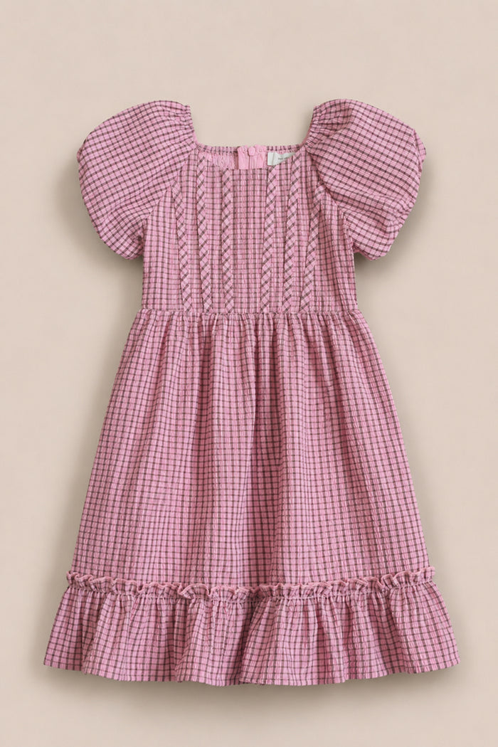 The Mini Monica Dress in Pink & Brown Gingham by Ivy City Co features short puff sleeves, a smocked bodice with decorative stitching, and a ruffled hem, capturing the classic charm of this girls’ style. Displayed on a neutral background.