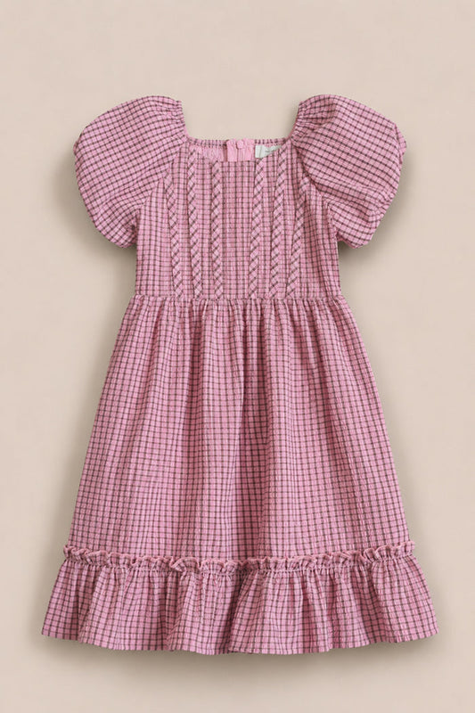 The Mini Monica Dress in Pink & Brown Gingham by Ivy City Co features short puff sleeves, a smocked bodice with decorative stitching, and a ruffled hem, capturing the classic charm of this girls’ style. Displayed on a neutral background.