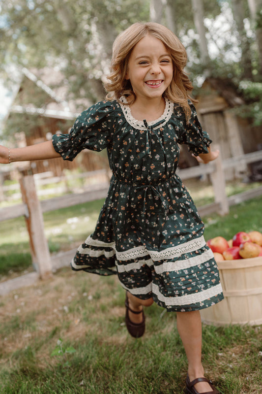 A young girl beams with joy as she runs outdoors near a wooden basket filled with apples, wearing the Ivy City Co Mini Palermo 3/4 Sleeves Dress in Evergreen Lovelorn Ditsy. Trees and rustic wooden buildings provide a charming backdrop.