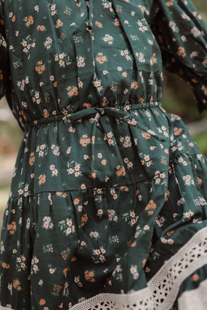 Close-up of the Ivy City Co Mini Palermo 3/4 Sleeves Dress in Evergreen Lovelorn Ditsy, showcasing its dark green shade with a delicate floral print, gathered waist, and white lace trim at the hem. This girls’ cotton dress features a slightly wrinkled texture for extra charm.