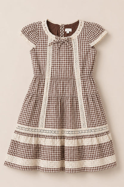 The Ivy City Co Mini Palermo Dress in Hickory Gingham features a brown and white gingham pattern, short puff sleeves, delicate lace trim, a round neckline with a small bow accent, and a tiered skirt crafted from a soft cotton-blend. The dress is shown laid flat on a neutral background.