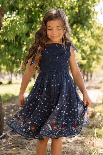 A young girl with long, wavy hair stands outdoors beneath a leafy tree on a sunny day, smiling as she holds the sides of her Ivy City Co Mini Rayna Dress in Midnight Cascading Floral. Sunlight filters through the branches, highlighting her airy chiffon dress—an ideal choice for fall festivities.