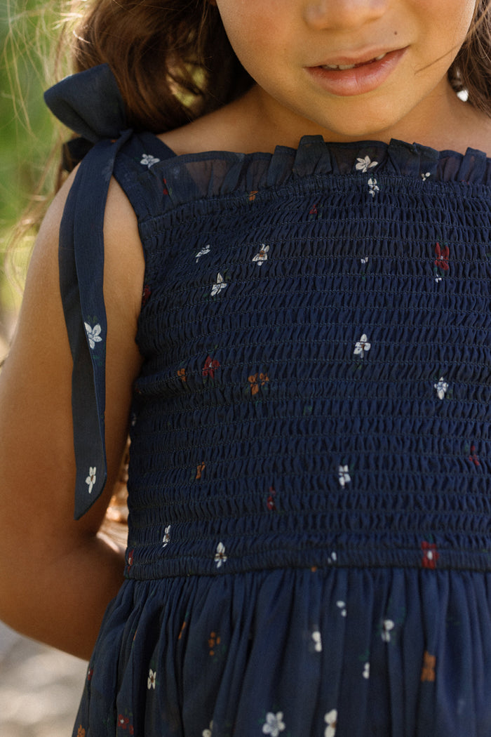 A young girl wearing the Ivy City Co Mini Rayna Dress in Midnight Cascading Floral—a navy blue smocked dress featuring delicate floral patterns and tied shoulder straps—stands outdoors, ready for fall festivities.