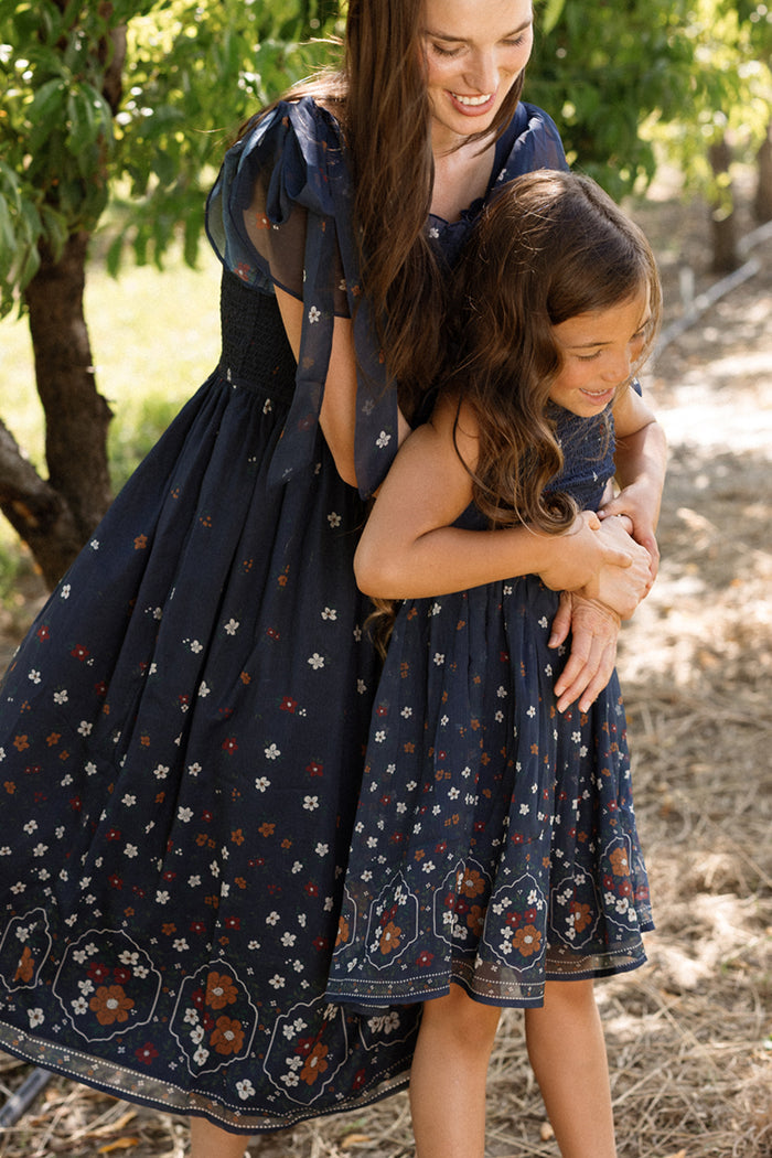A woman and a young girl, both wearing the Ivy City Co Mini Rayna Dress in Midnight Cascading Floral, smile and embrace outdoors near trees on a sunny day, their airy chiffon dresses adding a graceful touch to the moment.