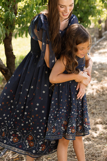 A woman and a young girl, both wearing the Ivy City Co Mini Rayna Dress in Midnight Cascading Floral, smile and embrace outdoors near trees on a sunny day, their airy chiffon dresses adding a graceful touch to the moment.