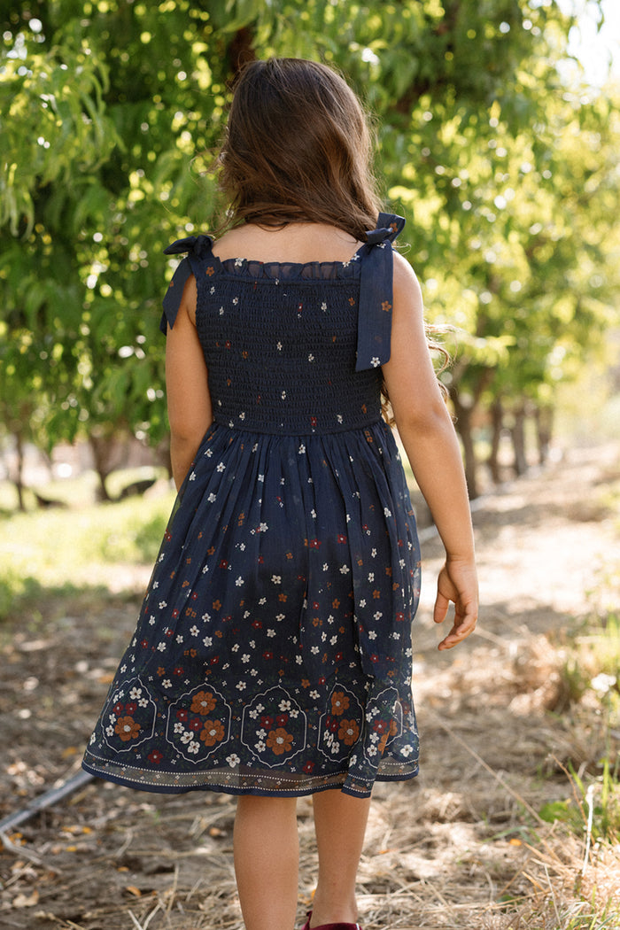 A young girl with long brown hair, wearing the airy Ivy City Co Mini Rayna Dress in Midnight Cascading Floral, walks away along a dirt path through a sunlit orchard lined with trees.