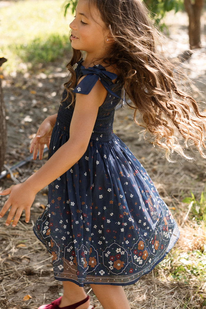 A young girl with long, wavy hair twirls outdoors on a sunny day, wearing the Ivy City Co Mini Rayna Dress in Midnight Cascading Floral. The airy chiffon dress features blue florals, and she is surrounded by trees, grass, and dappled sunlight.