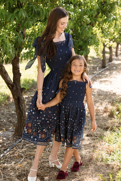 A woman and a young girl, both wearing matching Ivy City Co Mini Rayna Dresses in Midnight Cascading Floral, walk hand in hand through a sunlit orchard, smiling and enjoying the outdoors.