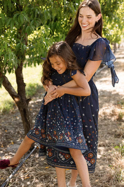 A smiling woman in the Ivy City Co Mini Rayna Dress in Midnight Cascading Floral hugs a laughing young girl wearing a matching airy chiffon dress outdoors near trees on a sunny day.
