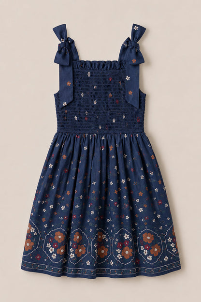The Mini Rayna Dress in Midnight Cascading Floral by Ivy City Co is a navy blue, sleeveless chiffon dress featuring a smocked bodice and bow-tied shoulder straps. Its skirt showcases larger floral patterns and geometric designs along the hem, creating the perfect airy look for fall festivities.