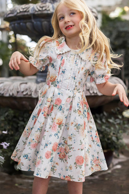 A young girl with long blonde hair wears the Mini Sophia Dress in Countryside Peony Blossom by Ivy City Co, standing outdoors, smiling and playfully raising her arms with greenery and a stone fountain in the background.