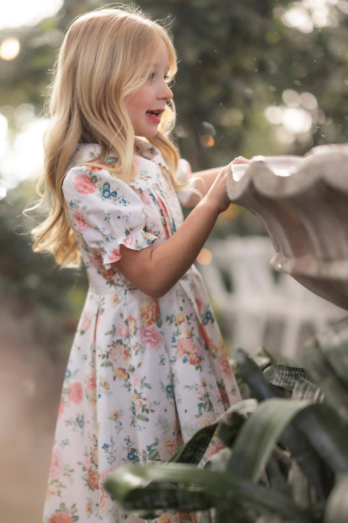 A young girl with long blonde hair wearing the Mini Sophia Dress in Countryside Peony Blossom by Ivy City Co stands by a stone fountain, gazing at it curiously, surrounded by lush greenery and soft sunlight.