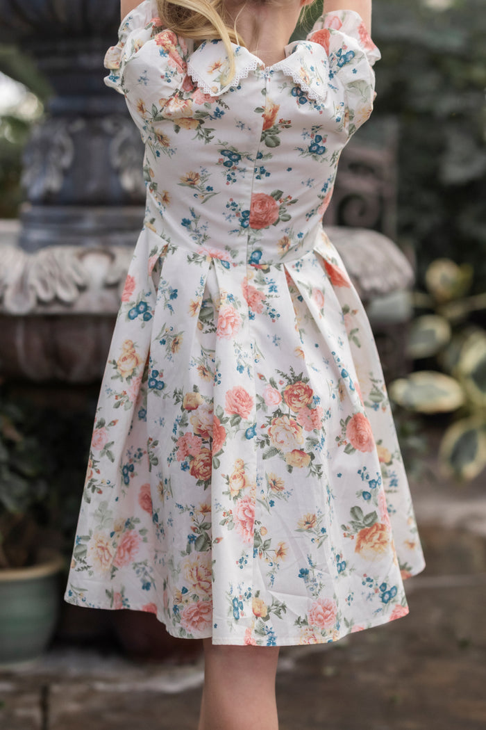 Wearing the Ivy City Co Mini Sophia Dress in Countryside Peony Blossom, a girl stands by a stone fountain outdoors, surrounded by lush greenery and potted plants.