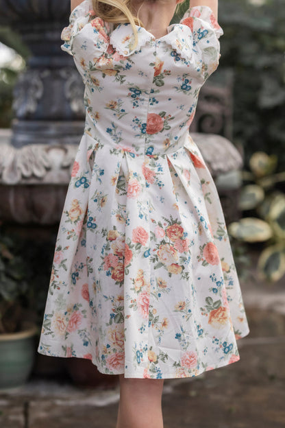 Wearing the Ivy City Co Mini Sophia Dress in Countryside Peony Blossom, a girl stands by a stone fountain outdoors, surrounded by lush greenery and potted plants.
