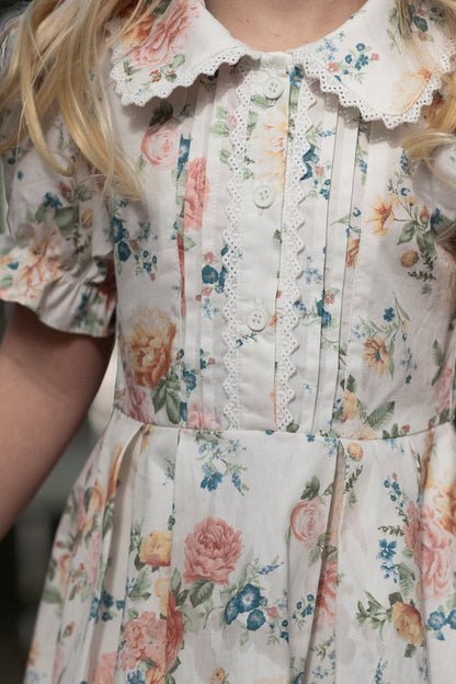 A close-up of a girl wearing the Ivy City Co Mini Sophia Dress in Countryside Peony Blossom, featuring an ivory floral print, lace-trimmed collar, pleated bodice, and white buttons. Only her upper body is visible.