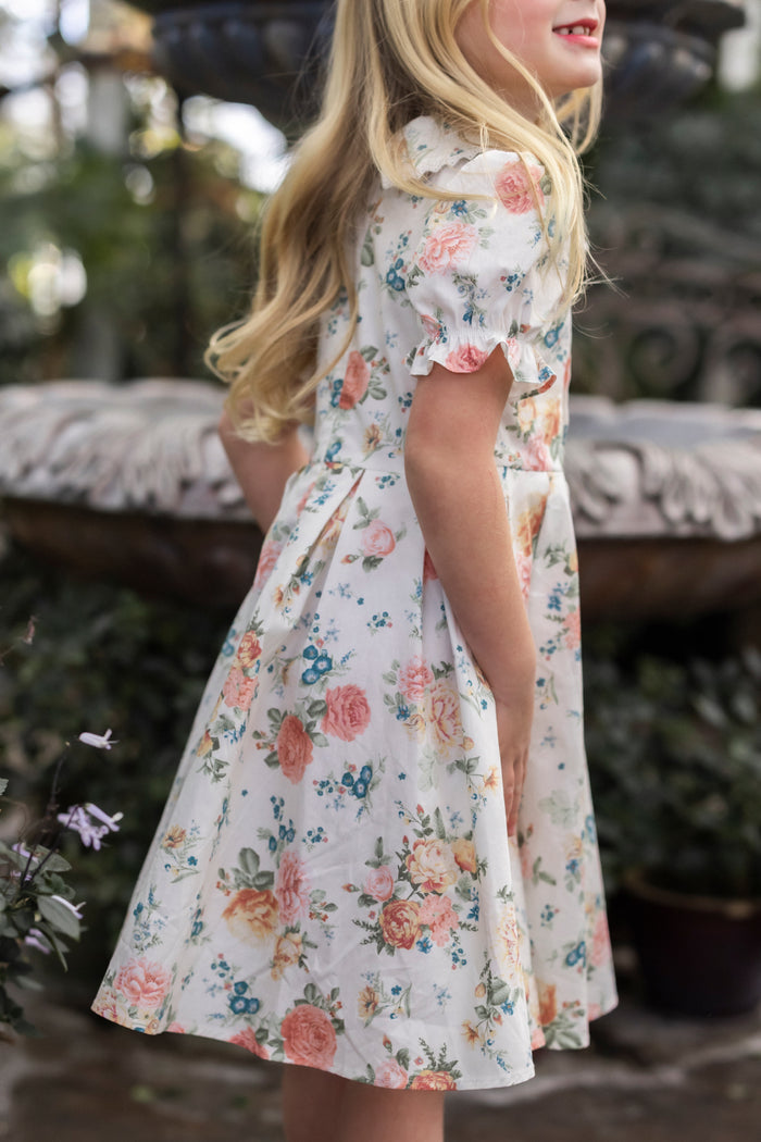A young girl with long blonde hair wears the Ivy City Co Mini Sophia Dress in Countryside Peony Blossom, standing sideways outdoors and smiling, surrounded by greenery and a decorative stone structure.