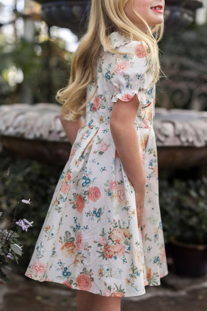 A young girl with long blonde hair wears the Ivy City Co Mini Sophia Dress in Countryside Peony Blossom, standing sideways outdoors and smiling, surrounded by greenery and a decorative stone structure.