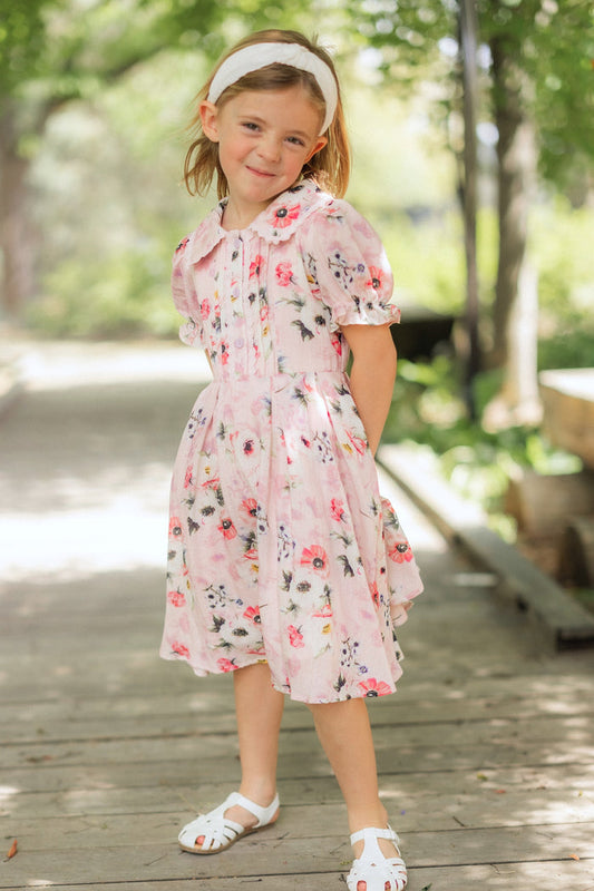 A young girl wears the Ivy City Co Mini Sophia Dress in Roselyn Pink with white sandals and a headband, standing on a wooden path and smiling amid green trees—perfect for those sweet mommy and me moments.
