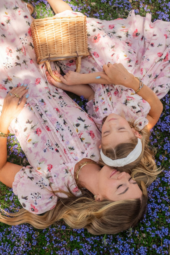 Two women in matching Ivy City Co Mini Sophia Dress in Roselyn Pink sets relax on grass with purple flowers, holding a wicker basket. Eyes closed, they enjoy the outdoors and sunlight together.