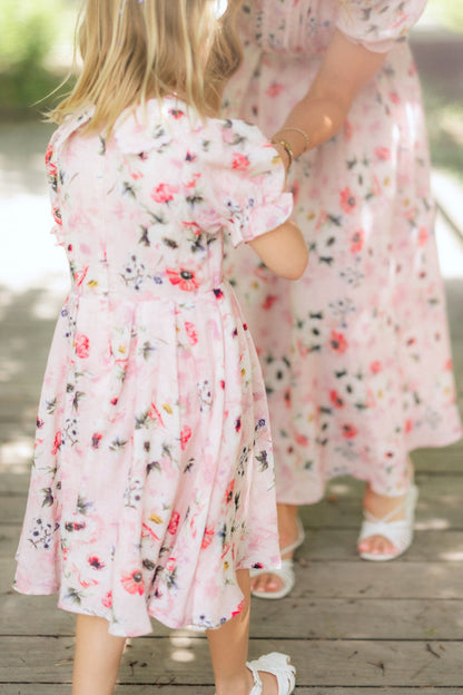 A young girl and a woman wear matching Ivy City Co Mini Sophia Dresses in Roselyn Pink with sandals on a wooden deck. The soft focus hints at a warm, sunny day.