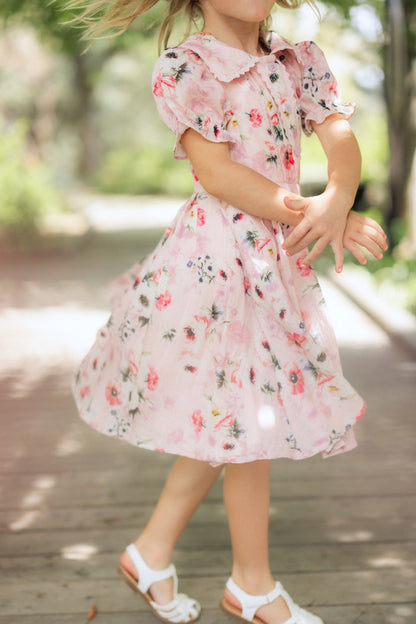 A young girl twirls joyfully on a wooden walkway, her blonde hair and Mini Sophia Dress in Roselyn Pink by Ivy City Co flowing, framed by a bright green outdoor backdrop—perfect for sunny days.