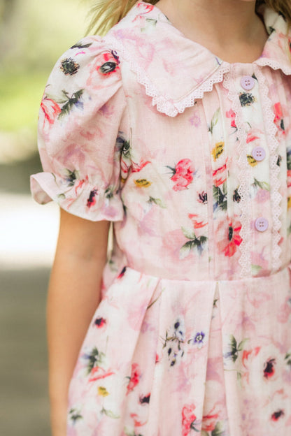 A close-up of a child in the Mini Sophia Dress in Roselyn Pink by Ivy City Co, featuring puffed short sleeves, lace-trimmed collar and front, and pink buttons. The softly blurred outdoor background highlights this charming dress.