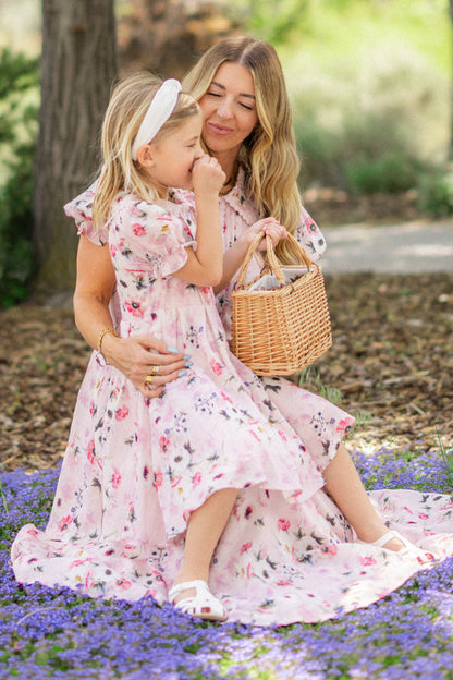 A woman and young girl wear matching Ivy City Co Mini Sophia Dresses in Roselyn Pink as they sit together on a field of purple flowers. The woman smiles, holding a wicker basket, while the girl pairs her dress with a white headband and sandals.