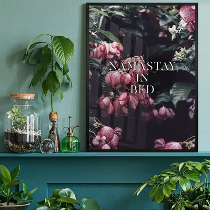 Namastay In Bed typography print in a black frame, featuring dark pink floral imagery on a teal shelf with indoor plants.