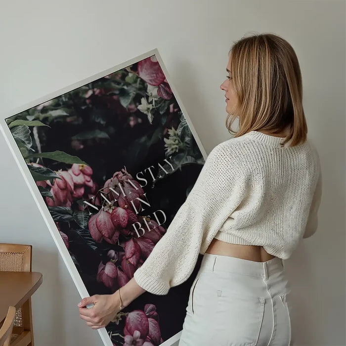 Woman holding a large Namastay In Bed typography print featuring pink floral botanical art in a white frame.