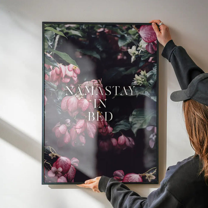 Namastay In Bed typography print in a black frame featuring pink flowers on a dark, moody botanical background.