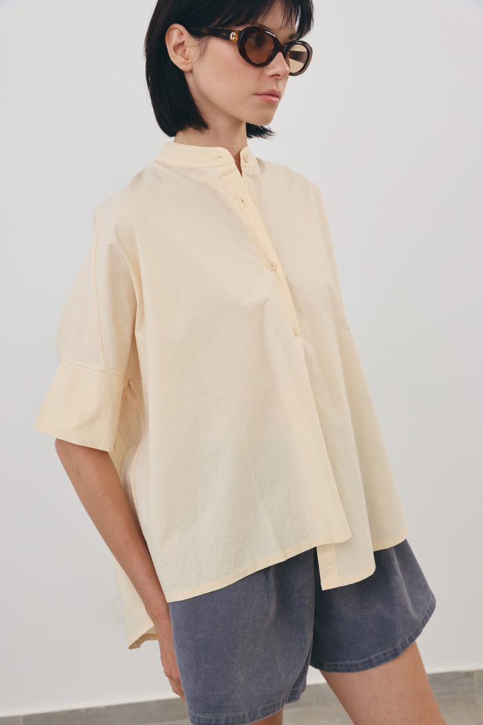 A woman wearing an oversized pink salmon-colored short-sleeve button-up shirt with a stand collar and dark gray denim shorts, photographed against a plain light gray wall.