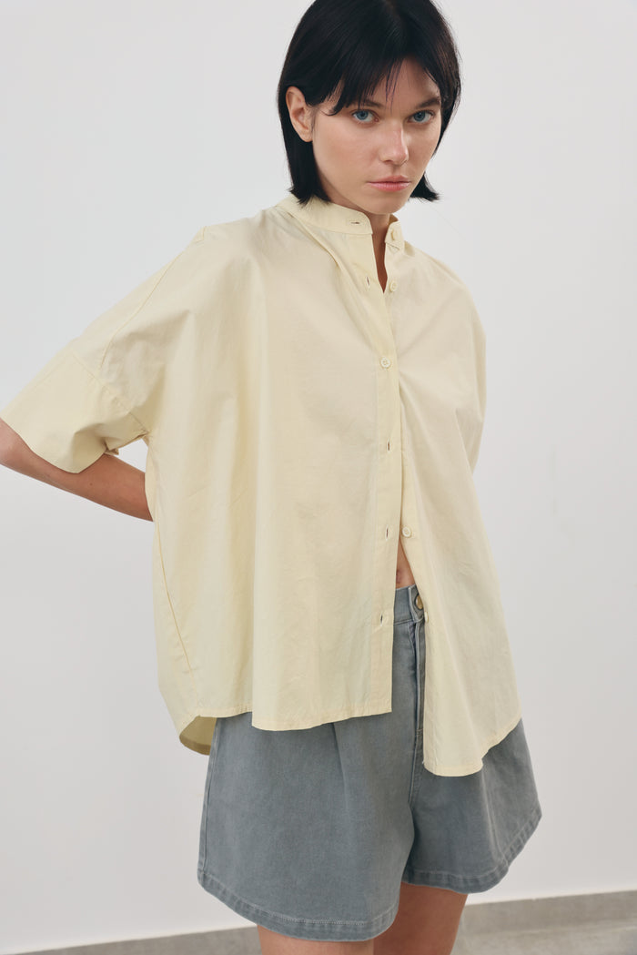 A model wearing an oversized, short-sleeved yellow sand-colored button-up shirt and gray denim shorts, standing against a plain light gray wall