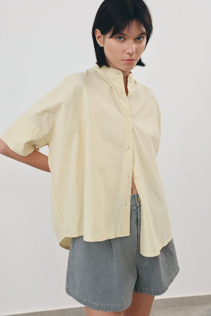 A model wearing an oversized, short-sleeved yellow sand-colored button-up shirt and gray denim shorts, standing against a plain light gray wall