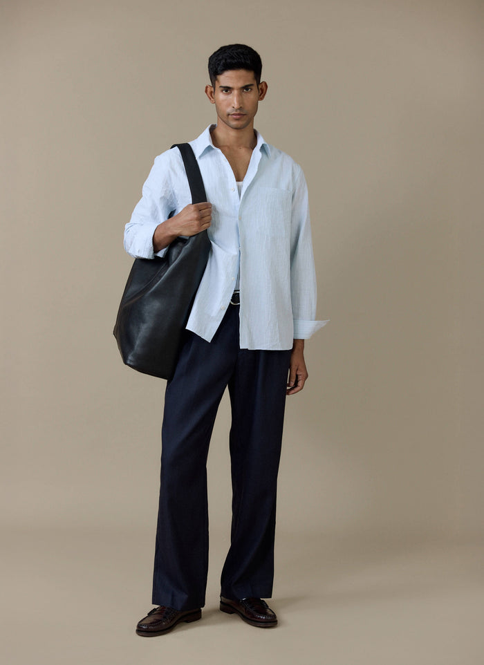 A model wearing the light blue Percival Paros Pearce Shirt in cotton seersucker with navy trousers and a black leather bag.