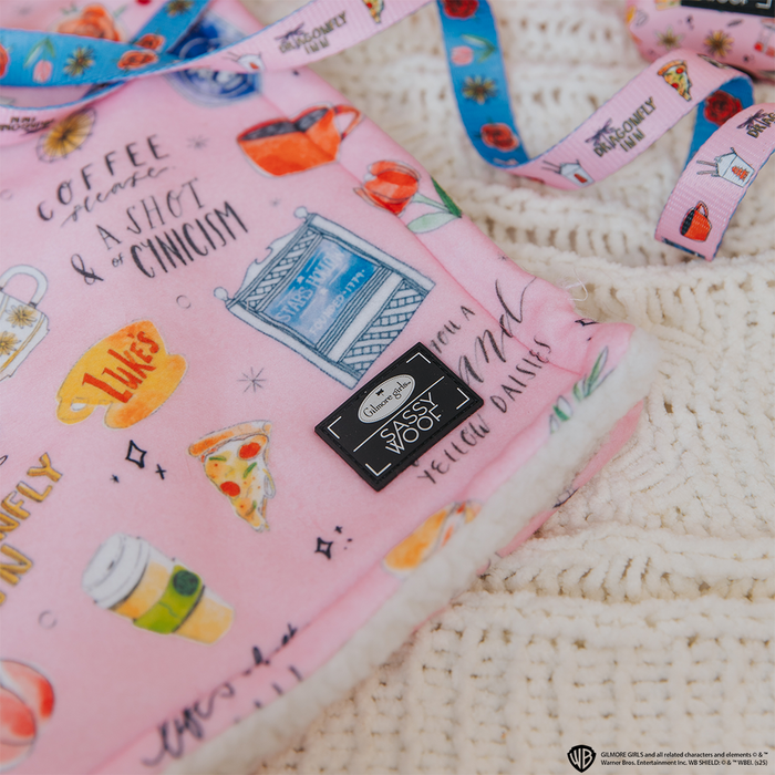 The Sassy Woof Pet Blanket - Gilmore Girls™ is a soft pink sherpa throw featuring playful illustrations of coffee cups, pizza, and witty text—perfect for any Stars Hollow fan. Shown here on a cream knit surface.