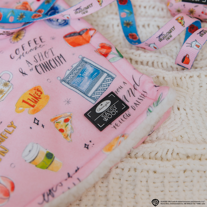 The Sassy Woof Pet Blanket - Gilmore Girls™ is a soft pink sherpa throw featuring playful illustrations of coffee cups, pizza, and witty text—perfect for any Stars Hollow fan. Shown here on a cream knit surface.