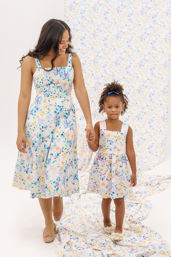A woman and a young girl hold hands, both wearing the Ivy City Co Petal Dress in Blue Bouquet. The background displays matching floral fabric. The woman smiles at the girl as she looks toward the camera.