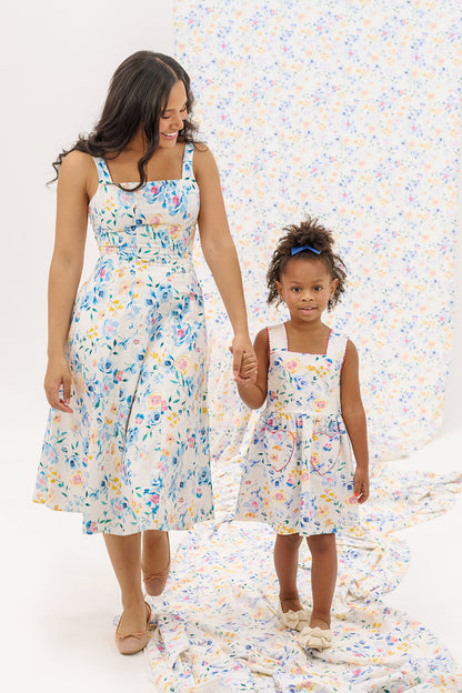 A woman and a young girl hold hands, both wearing the Ivy City Co Petal Dress in Blue Bouquet. The background displays matching floral fabric. The woman smiles at the girl as she looks toward the camera.