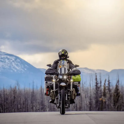 Person riding a motorcycle with luggage and a Primus fuel bottle against a mountainous landscape