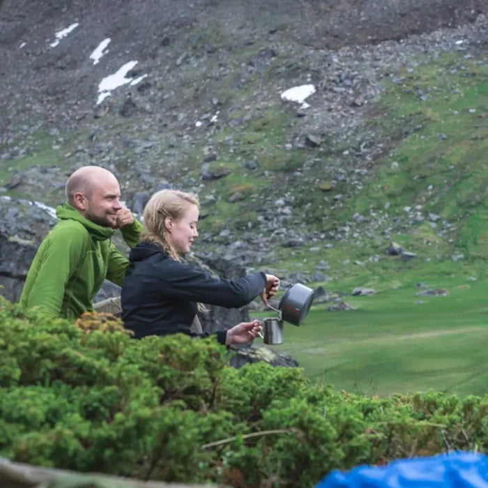 Two people sitting outdoors in a mountainous area, one pouring a drink from a Primus Litech 1500ml camp kettle.